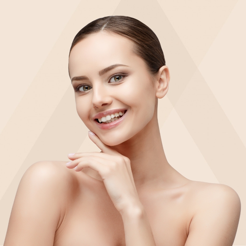 A young woman with smooth, glowing skin is smiling brightly, showing her teeth. She has neatly groomed eyebrows, subtle makeup, and her dark hair is pulled back into a sleek style. Her hand is gently touching her chin, and she is posed against a soft beige background with faint geometric patterns.